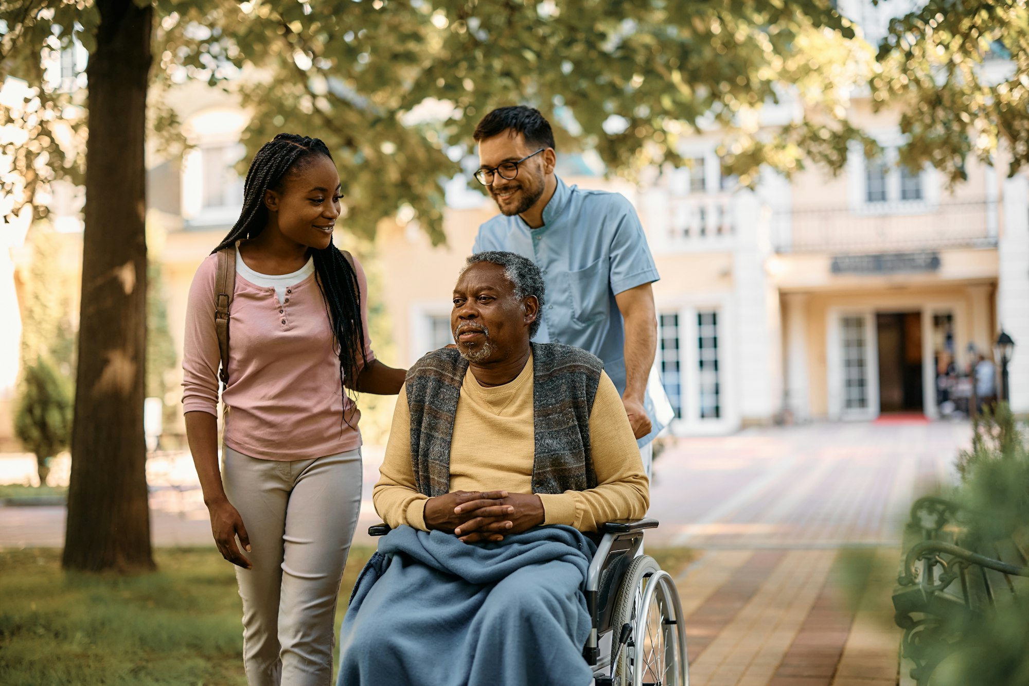 Black senior in wheelchair enjoying with his daughter and caregiver in the park at nursing home.
